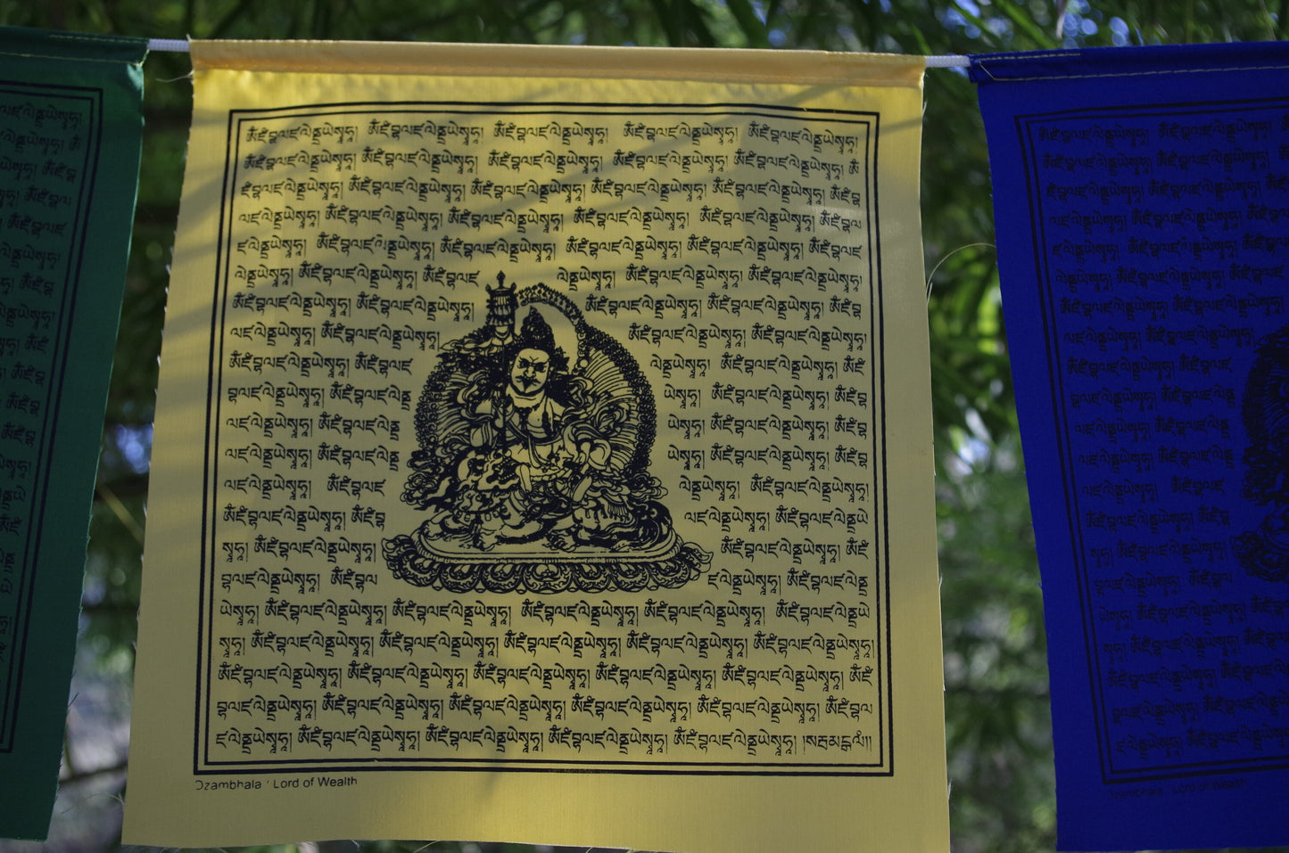 one yellow 10 inch by 10 inch Dzambhala wealth deity prayer flag from a five color set hanging outside under bamboo