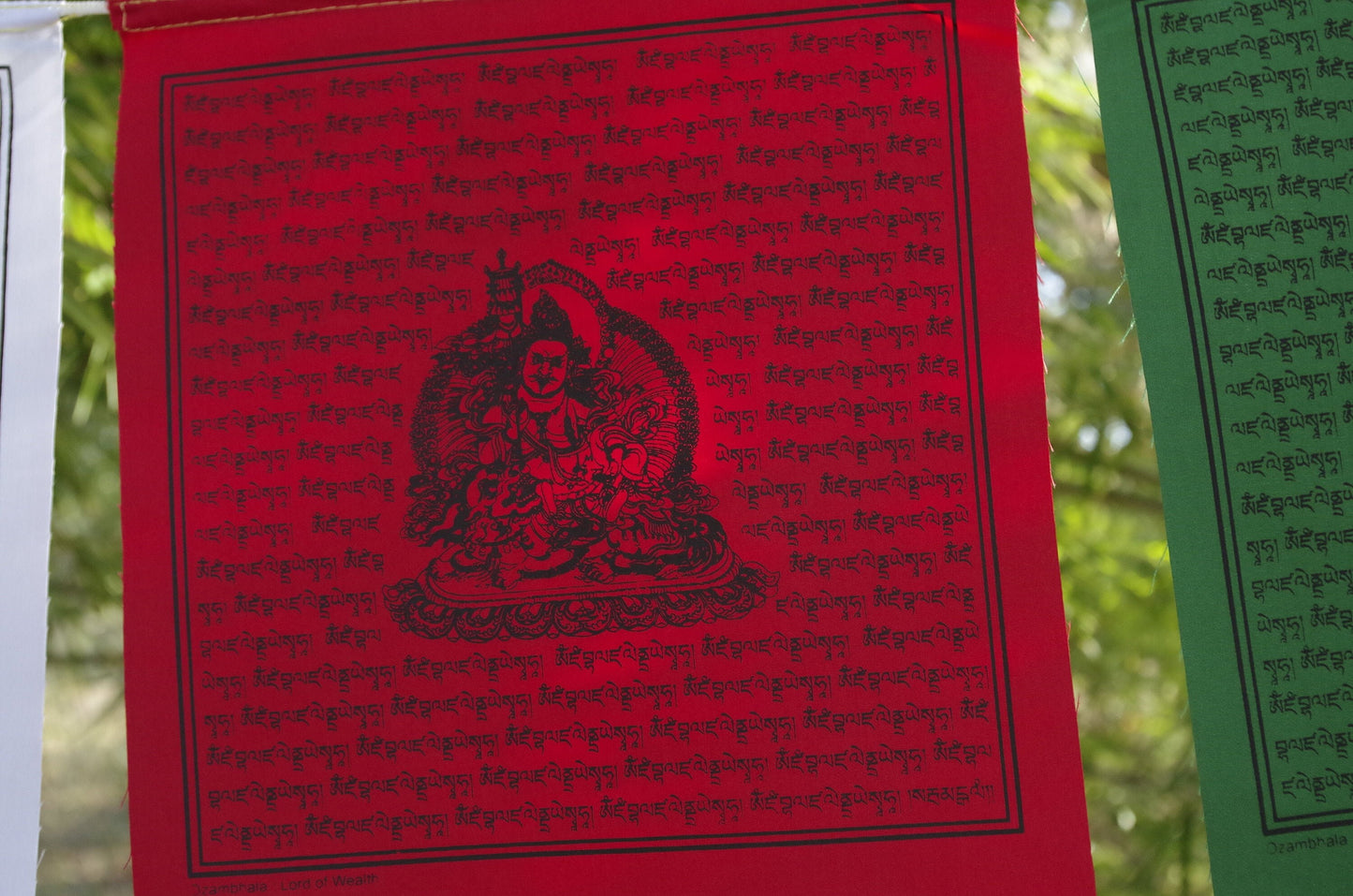 one red 10 inch by 10 inch Dzambhala wealth deity prayer flag from a five color set hanging outside under bamboo