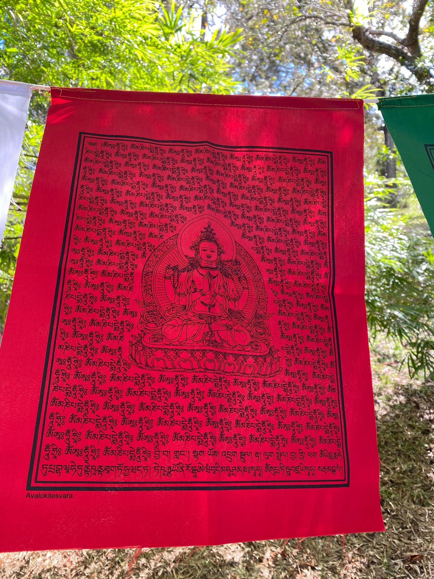 A close up of 1 red Chenrezig prayer flag in 5 colors, each 14x17 inches, depicting the Buddha of Compassion hanging outdoors.