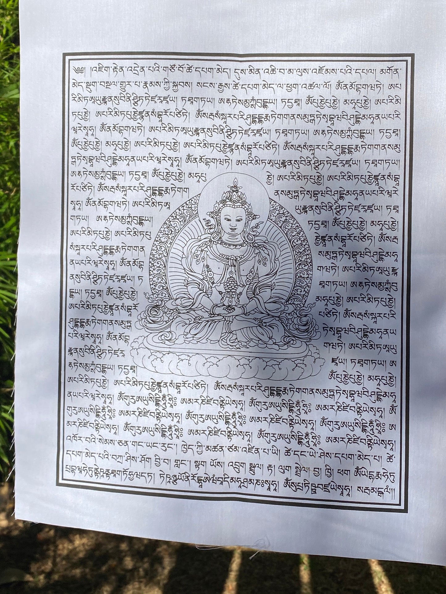 A close up of a 14&quot;x17&quot; white Amitayus prayer flag, featuring The Buddha of Boundless Life, hanging in dappled sunlight for the spreading of abundant long life blessings in every direction