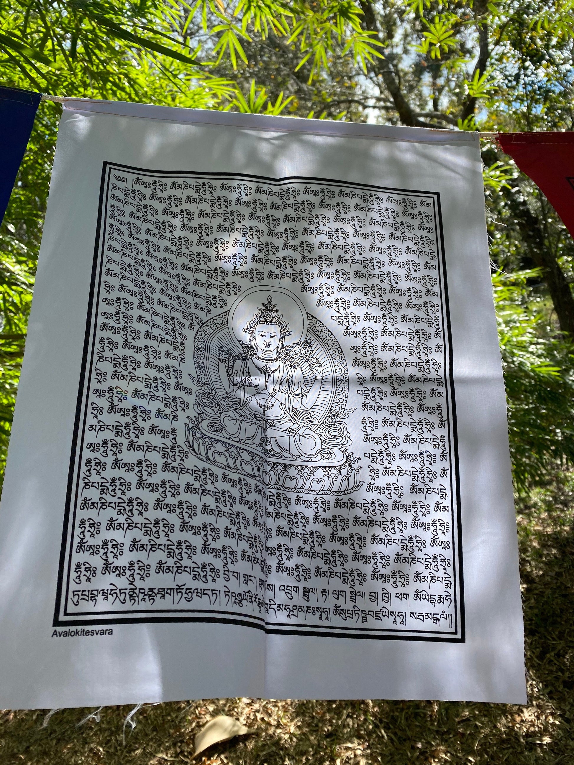 A close up of 1 white Chenrezig prayer flag in 5 colors, each 14x17 inches, depicting the Buddha of Compassion hanging outdoors.