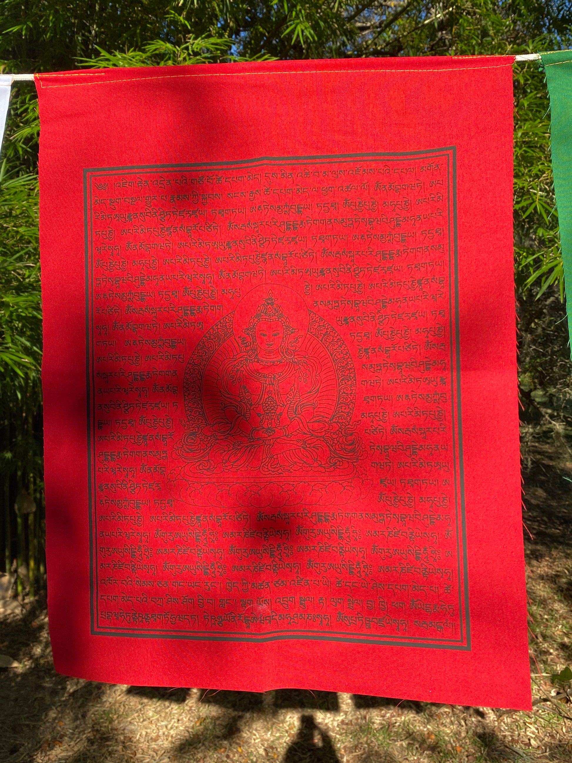 A 14&quot;x17&quot; red Amitayus prayer flag, featuring The Buddha of Boundless Life, hanging in dappled sunlight for the spreading of abundant long life blessings in every direction