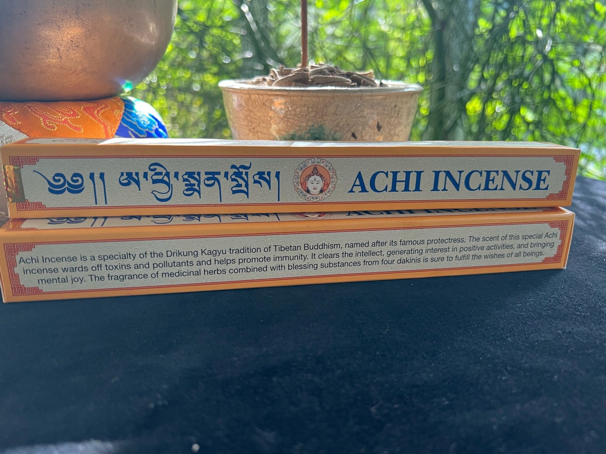 Close-up of Achi Incense boxes with Tibetan and English product descriptions on black velvet, accompanied by a 5 metal sound bowl and raku incense bowl. Experience the benefits of our specialty incense for your meditation practice.