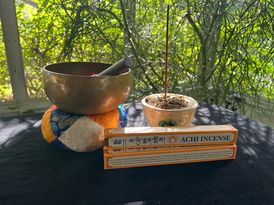 Stack of yellow and white boxes of Achi  Incense on black velvet, accompanied by a 5 metal sound bowl and a raku fired incense bowl with a stick of Achi Incense. Enhance your meditation experience with our specialty incense.