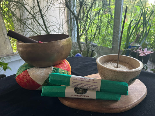 Rolls of Himalayan Arts Juniper Incense on a table with one of the sticks in an incense burner waiting to be lit. The incense is wrapped in green paper.  Also on the table is a Tibetan singing bowl.